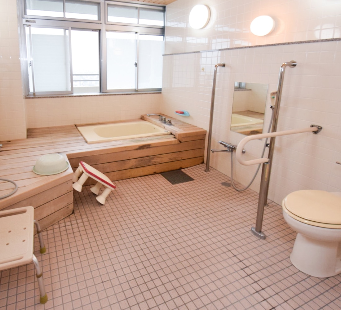 木製の浴槽と手摺り、トイレが備わった介護施設の浴室。採光良好で安全設計。